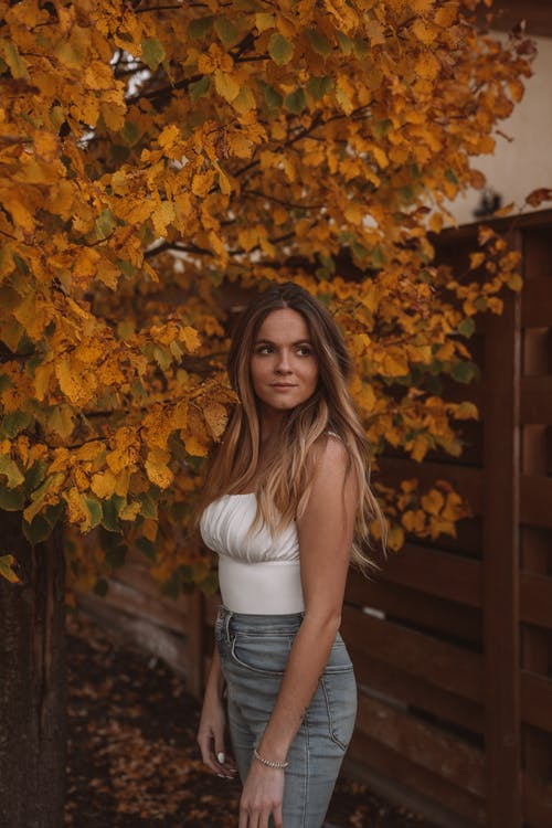 A Woman in a White Tank Top and Jeans Standing Under an Autumn Tree