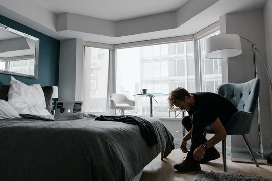 An Unmade Made with a Laptop on it, and a Man Sitting on a Chair, and Tying His Shoelaces, Looking Ready to Leave
