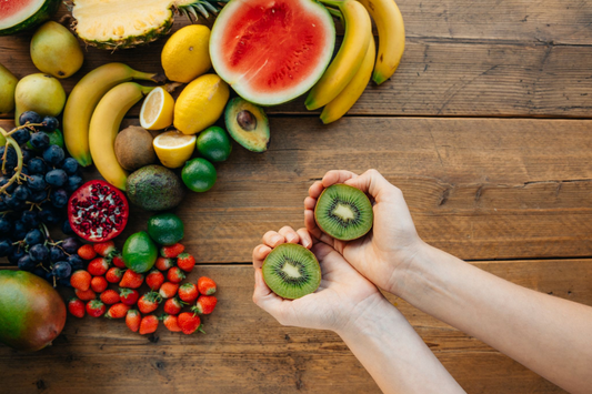 A person holding kiwi and many other fruits laying on a wooden table