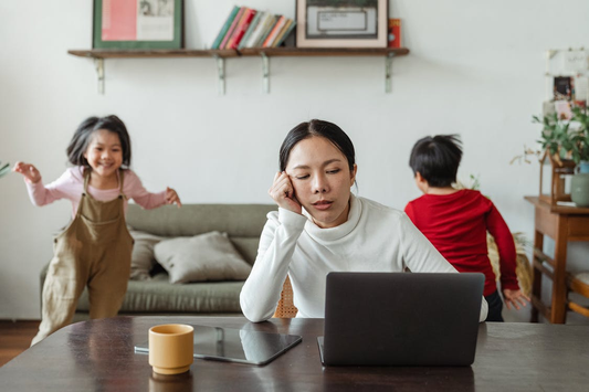 A Female Sitting in Front of a Laptop, Looking Visibly Distressed, and Two Kids Running in the Background