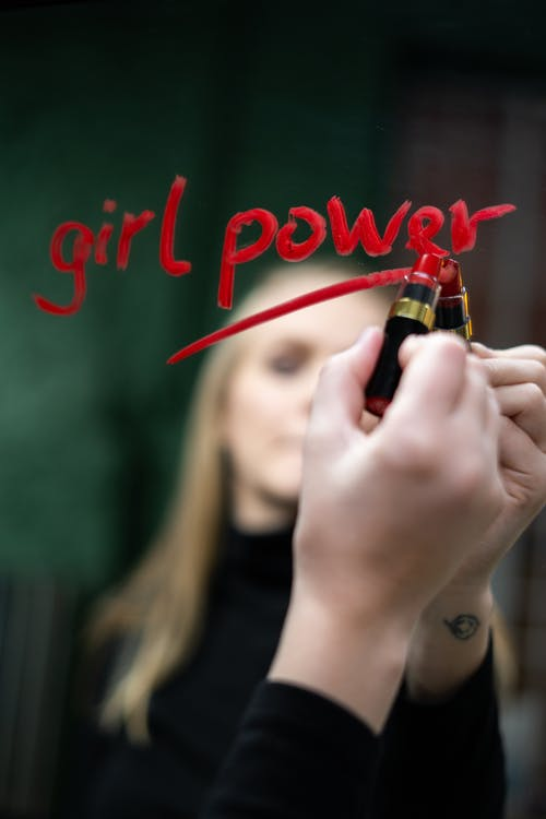 A Blonde Female Writing “Girl Power” on a Mirror with Red Lipstick