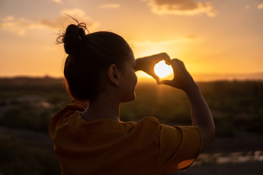 A woman making a heart sign with her hands in front of the sun.